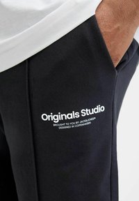 Pantalons de survêtement noirs en coton avec un imprimé logo blanc qui dit "Originals Studio" sur la cuisse gauche, comportant une poche latérale.