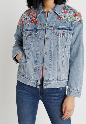 Lichtblauwe spijkerjas met kleurrijke bloemenborduursels op de schouders, gedragen over een spijkerbroek, handen van het model gedeeltelijk in de zakken.
