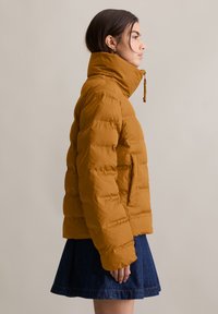 Mosterdgeel pufferjack met een hoge kraag en zijzak, voorzien van een doorgestikt ontwerp en een gladde afwerking, gecombineerd met een jeansrok.