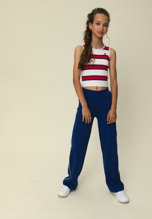 Tienermeisje met lang gevlochten haar, gekleed in een witte cropped top met rode strepen, marineblauwe wijde broekspijpen en witte sneakers, staand tegen een neutrale achtergrond.