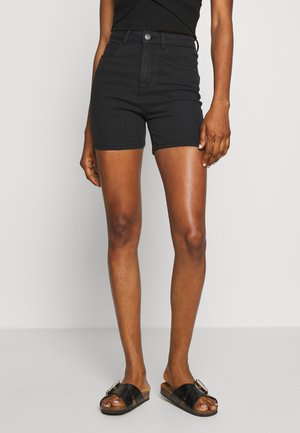 Vrouw draagt zwarte high-waist denim shorts en zwarte slides met gesp, staand tegen een effen lichte achtergrond.