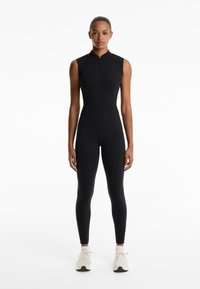 Zwarte mouwloze jumpsuit met een hoge kraag en ritssluiting aan de voorkant, aansluitend ontwerp met een gladde textuur, gecombineerd met lichte sportschoenen.