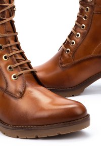 Stivali ankle in pelle marrone con finitura liscia, design con lacci, occhielli dorati e suola in gomma con scanalature di trazione.