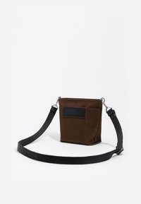 Borsa in tessuto marrone di forma rettangolare, con un'etichetta nera sul fronte, una tracolla staccabile e dettagli in metallo.