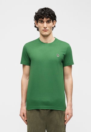 Νέος άντρας με σγουρά μαλλιά, φορώντας ένα απλό πράσινο T-shirt με στρογγυλή λαιμόκοψη και ένα μικρό έμβλημα ζώου στο στήθος, στέκεται με χαλαρά δάχτυλα.