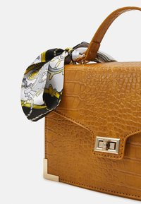 Sac à main en cuir gaufré couleur fauve avec fermoir doré et détails aux coins, orné d'un foulard à motifs noir, blanc et jaune attaché à la poignée.