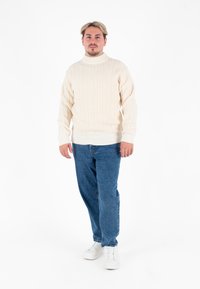 Flauschiger cremefarbener Rollkragenpullover mit gerippter Textur, kombiniert mit blauen Jeans und weißen Sneakern. Schlichte, lockere Passform.