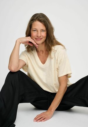 Camicia crema a maniche corte con scollo a V, realizzata in tessuto leggero, abbinata a pantaloni neri larghi. Il modello è seduto con le mani poggiate sulle ginocchia.