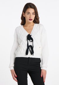 Weißer Strickcardigan mit Rautenmuster, V-Ausschnitt, schwarzen Bändern an der Vorderseite und zwei seitlichen Taschen mit Knöpfen.