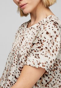 Blusa beige con motivo a pois marroni, maniche corte e texture morbida. Presenta un scollo a V e una vestibilità leggermente ampia.