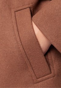 Main partiellement insérée dans une poche latérale rectangulaire d'un manteau en laine marron avec des coutures visibles.