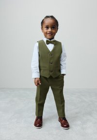 Jeune enfant debout, portant un gilet et un pantalon vert olive, une chemise blanche, un nœud papillon assorti et des chaussures habillées marron sur un sol gris.