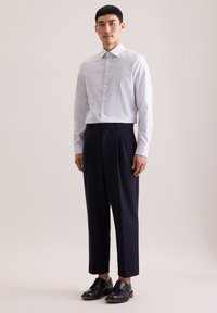 Camicia bianca con bottoni e pois blu, abbinata a pantaloni plissettati blu navy e scarpe di pelle nere. Tessuto liscio e design classico.