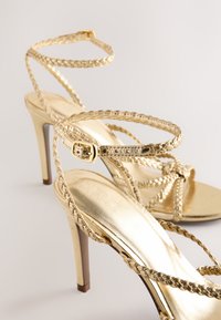 Sandales à talons hauts dorées avec lanières tressées, finition métallique et design élégant. Comprend une boucle réglable et une semelle intérieure texturée.