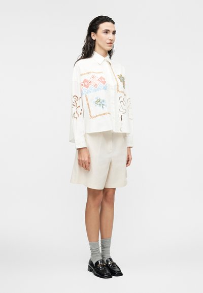 Γυναίκα με σκούρα μαλλιά που φοράει άνετο λευκό πουκάμισο με πολύχρωμη κεντήματα, κρεμ σορτς μέχρι το γόνατο, γκρι κάλτσες και μαύρα μοκασίνια, στέκεται πλαγίως.