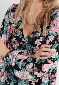 Kukallinen mekko, jossa on musta tausta ja kirkkaan pinkkejä, turkooseja ja valkoisia kukkia. Siinä on pitkät hihat ja V-pääntie.