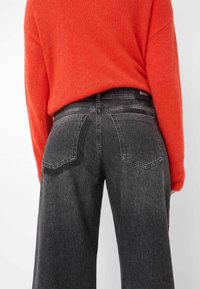 Schwarze weit geschnittene Jeans mit ausgewaschenem Waschung und Gesäßtaschen, kombiniert mit einem orangefarbenen strukturierten Pullover. Sichtbares Markenlogo am Bund.