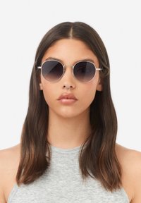 Ringikujulised päikeseprillid tumedate gradienteedidega läätsede, õhukese hõbedase metallraami ja nina silla aktsendiga. Kandja on keskmise pikkusega juustega inimene.