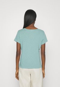 Tričko s krátkým rukávem v jemném tyrkysovém materiálu, volný střih, hladká textura, kulatý výstřih, s zakřiveným lemem a detailním prošitím na bocích.