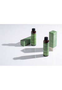 Två mörk amberfärgade flaskor med toner, med gröna etiketter, bredvid matchande gröna och vita förpackningar på en vit yta, som kastar långa skuggor.