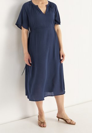 Frau trägt ein marineblaues Midikleid mit Schmetterlingsärmeln und V-Ausschnitt, kombiniert mit braunen Sandalen mit niedrigem Absatz, steht vor einer weißen Wand.