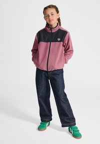 Fleecejack in roze en zwart, met ritssluiting, voorzien van een hoge kraag en zijzakken, gecombineerd met een donkere spijkerbroek en groene sneakers.