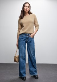 Beżowy puszysty sweter z rękawami o długości trzech czwartych, zestawiony z niebieskimi spodniami dzwonami z wysokim stanem. Modelka trzyma małą beżową torebkę.