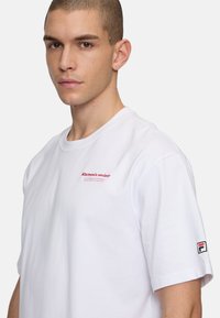 Hvid bomulds t-shirt med korte ærmer, der har rød tekst på brystet og et farvet logo-lap på venstre ærme. Glat tekstur.