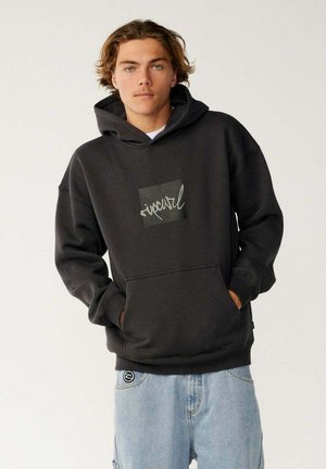 Młody mężczyzna o jasnobrązowych włosach, ubrany w czarną bluzę z kapturem Rip Curl i jasnoniebieskie dżinsy, trzymający ręce w kieszeniach bluzy.