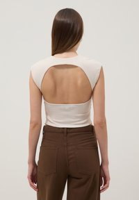 Mujer con cabello largo y castaño que lleva una blusa sin mangas color crema con la espalda descubierta y pantalones marrones de talle alto, de pie frente a un fondo liso.