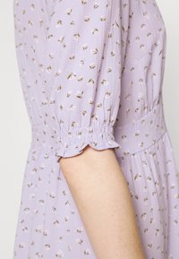 Robe lavande avec des manches courtes et bouffantes présentant un motif floral en rose et vert. Tissu texturé avec des poignets de manches élastiqués et une taille élastique.