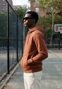 Jeune homme portant des lunettes de soleil, un sweat à capuche marron et un pantalon blanc, debout les mains dans les poches sur un terrain de basket en plein air près d'une clôture.