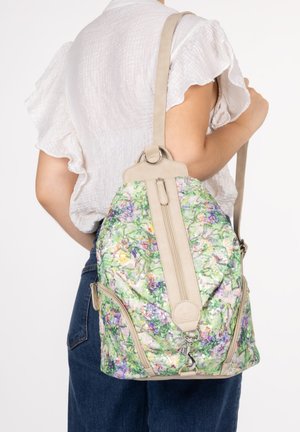Woman wearing white ruffled sleeve blouse and blue jeans carrying floral patterned green and purple backpack with beige straps over shoulder.