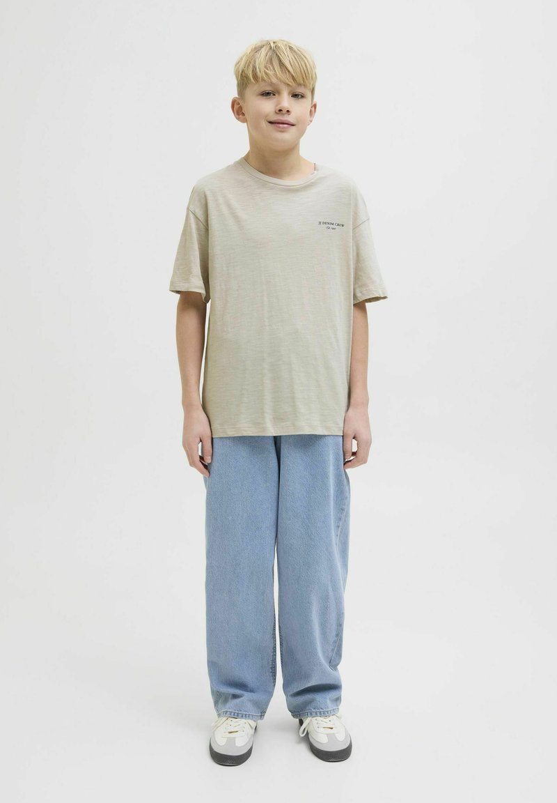 Beige T-shirt met korte mouwen en een bedrukt logo, gecombineerd met losse lichtblauwe denim broek en grijs-witte sneakers. Eenvoudige witte achtergrond.