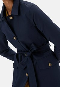 Granatowy płaszcz trencz wykonany z gładkiej tkaniny, z paskiem w talii, zapięciami na guziki oraz dwoma przednimi kieszeniami z zaokrąglonymi mosiężnymi guzikami.