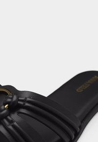 Μαύρο σανδάλι με σαγιονάρα με πλεκτούς δερμάτινους ιμάντες και χρυσή κυκλική διακοσμητική λεπτομέρεια στην κορυφή, με λεία εσωτερική σόλα και διακριτικό ανάγλυφο λογότυπο.