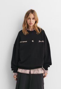 Čierna oversized mikina s nápisom "FULLY SUPPORT" a dekoratívnou výšivkou. Nosená cez ružovú kockovanú košeľu, s dlhými rukávmi a rebrovanými manžetami.