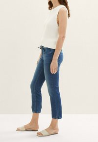 Mujer con una blusa de punto blanca sin mangas, jeans cortos azules y sandalias beige de plié, de pie contra un fondo claro y liso.