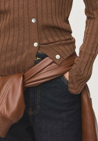 Cardigan a coste marrone con bottoni argentati, abbinato a pantaloni in denim scuri e una cintura in pelle marrone annodata in vita.