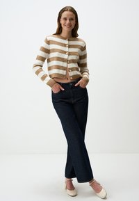 Geknipte cardigan in crèmekleur met tan strepen en knoopsluiting. Hoog-taille donkere blauwe jeans. Model draagt crèmekleurige hakken en staat in een neutrale achtergrond.