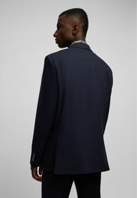 Marineblauwe gestructureerde blazer met een enkele vent en knoopmanchetten, gedragen door een model gezien vanaf de achterkant.