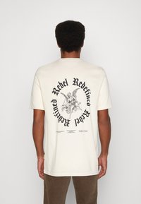 Cremfarbendes Baumwoll-T-Shirt mit einem großen schwarzen Grafikmotiv auf der Rückseite, das den Text "Rebel Redefined" und eine Engel-Illustration zeigt.