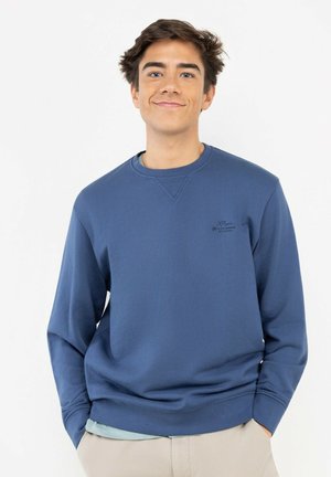 Joven con sudadera azul y pantalones beige, de pie con las manos en los bolsillos, sonriendo ligeramente sobre un fondo blanco liso.