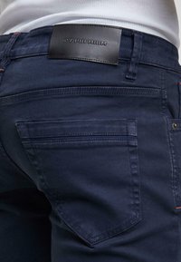 Tmavě modré džíny z denimu s hladkou texturou, standardními kapsami a obdélníkovou černou koženou nášivkou s nápisem "2Y PREMIUM" na pase.