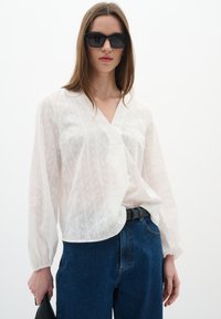 Blusa bianca testurizzata con scollo a V e maniche lunghe, abbinata a jeans in denim blu a vita alta. Occhiali da sole neri e una borsa nera completano il look.