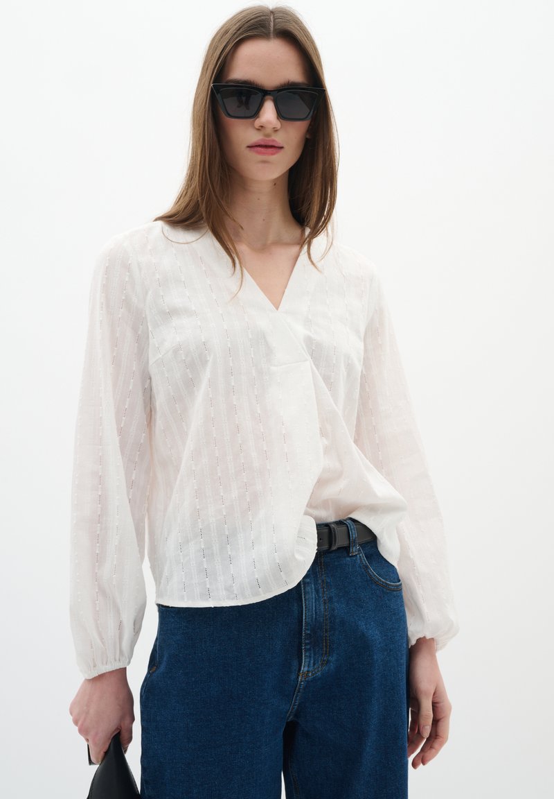 Blusa bianca testurizzata con scollo a V e maniche lunghe, abbinata a jeans in denim blu a vita alta. Occhiali da sole neri e una borsa nera completano il look.