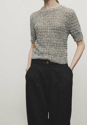 Femme portant un haut tricoté à manches courtes texturé dans des tons gris, rentré dans un pantalon noir taille haute à jambes larges, les mains dans les poches.