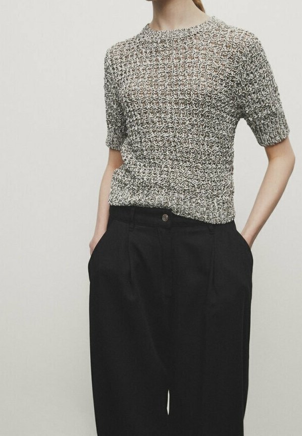 Femme portant un haut tricoté à manches courtes texturé dans des tons gris, rentré dans un pantalon noir taille haute à jambes larges, les mains dans les poches.