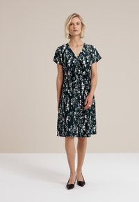 Modèle dans une robe à motifs, longueur genou, à manches courtes, se tient sur un fond clair, portant des chaussures noires. Apparence élégante et posée.