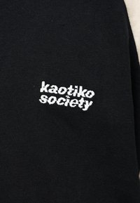 Svart stoff med hvit brodert tekst "kaotiko society" sentrert på det synlige området av materialet.